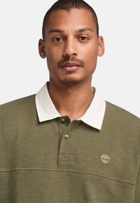 Olivgrön polo skjorta med vit krage, med en subtil trälogotyp på bröstet och en strukturerad tygfinish. Tre-knapparsplacket.