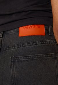 Svarte denimbukser med en teksturert overflate og en oransje lærpatch med "beWider" utformet på baksiden av midjebåndet.