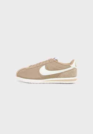 Baskets Nike Cortez beiges avec logo Swoosh blanc, semelle blanche et accents en daim, présentées sur un fond clair uni.