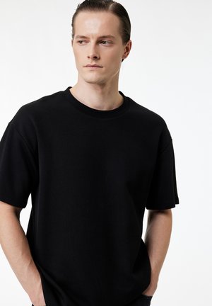 Jonge man met naar achteren gestyled haar, die een eenvoudige zwarte korte mouw T-shirt draagt, kijkt opzij met handen in de zakken.