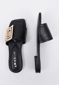 Chanclas negras con una tira ancha que presenta una hebilla cuadrada dorada. Material sintético liso, suela plana y diseño minimalista.
