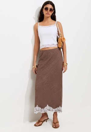 Femme portant un débardeur blanc orné de dentelle, une longue jupe marron avec un ourlet en dentelle blanche, des lunettes de soleil, des sandales, des bracelets en or et un petit sac à bandoulière.