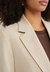 Beige linned blazer med skarpe revers, der har et tekstureret stof. En mørkebrun top er synlig under, hvilket giver en farvekontast.