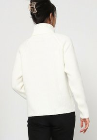 Pull en maille côtelée blanc avec un col montant, des manches longues et une coupe décontractée. La texture semble douce et chaude, adaptée aux temps frais.