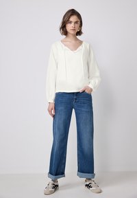Hvid pullover med snor og lange, puffede ærmer, parret med højtaljede, brede jeans i blå, rullet op ved manchetterne.
