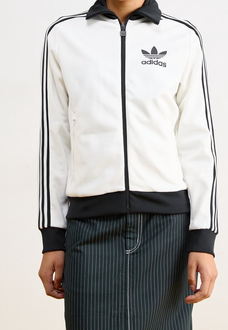 Giacca sportiva bianca con accenti neri, chiusura frontale con zip e l'iconico logo Adidas. Presenta strisce nere lungo le maniche, tessuto liscio.
