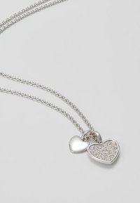 Collier en chaîne argentée avec deux pendentifs en forme de cœur, l'un lisse et l'autre incrusté de petits cristaux clairs.