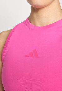 Rožinė sportinė tankų palaidinė su glaudžiu dizainu. Yra tekstūruota medžiaga ir subtilus iškilus logotipas, atitikantis tris įstrižas juosteles.