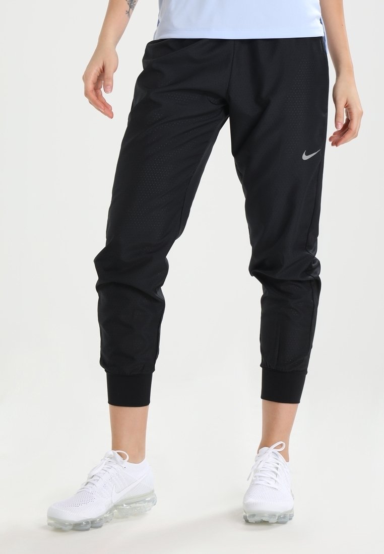 Zwarte sportbroek met een gestructureerd oppervlak, cropped ontwerp, elastische boorden en een klein zilveren logo aan de linkerkant; gecombineerd met witte sneakers.
