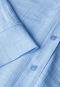 Camicia di tessuto blu chiaro sgualcita con colletto pieghevole e chiusura con bottoni dotata di un bottone arrotondato. Finitura morbida e design informale.