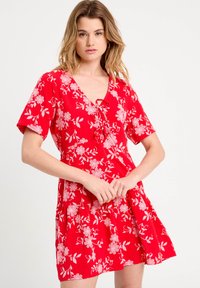 Robe à motif floral rouge avec un décolleté en V, des manches courtes et une coupe décontractée avec une jupe froncée. Le design inclut des accents floraux blancs.