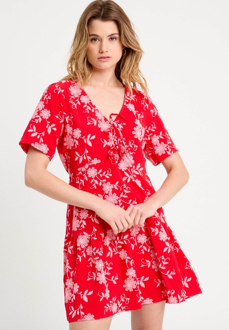 Robe à motif floral rouge avec un décolleté en V, des manches courtes et une coupe décontractée avec une jupe froncée. Le design inclut des accents floraux blancs.