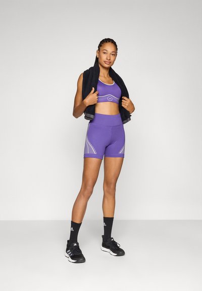 Lila sportmelltartó és rövidnadrág szett fényvisszaverő részletekkel. A modell egy fekete törölközőt tart a kezében. Az összeállítást fekete sportzokni és cipő egészíti ki.