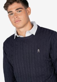 Granatowy sweter dzianinowy z teksturowanym wzorem w warkocze, posiadający ściągacz przy okrągłym dekolcie i małe haftowane logo na piersi.
