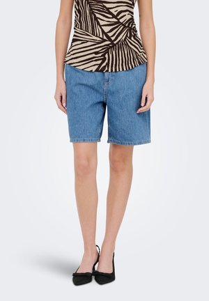 Femeie care poartă pantaloni scurți din denim albastru de lungime medie, un top cu model zebrat și tocuri negre ascuțite cu un mic fundiță.