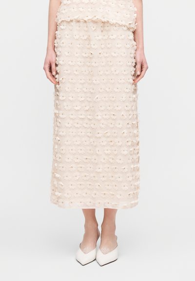 Jupe beige ornée de fleurs en tissu transparent, avec des motifs floraux en 3D, longueur genou, associée à des talons blancs à bout pointu.