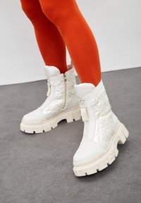 Legs in bright orange tights wearing white quilted ankle boots with chunky soles and front zippers on gray floor.