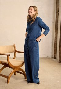 Femme souriante en chemise en denim bleu et pantalon à jambes larges, debout les mains dans les poches à côté d'une chaise en bois avec un coussin beige dans une pièce minimaliste.