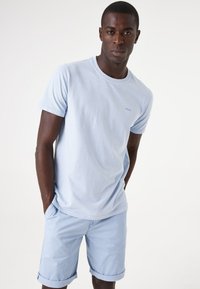 Lichtblauwe katoenen t-shirt met korte mouwen, ronde hals, en een klein logo op de borst, gecombineerd met lichtblauwe shorts met opgerolde mouwen.