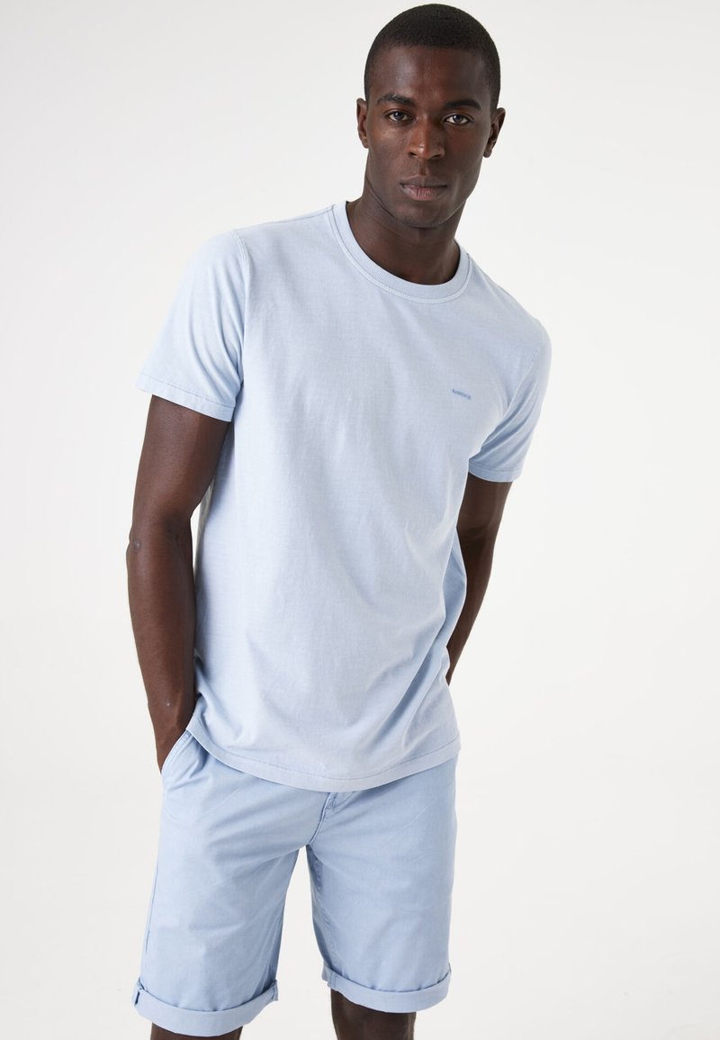 Lichtblauwe katoenen t-shirt met korte mouwen, ronde hals, en een klein logo op de borst, gecombineerd met lichtblauwe shorts met opgerolde mouwen.