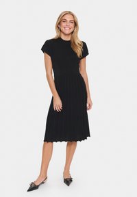 Black dress with short sleeves, textured bodice, flowing pleated skirt, and scalloped hem. Worn with black pointed-toe flats.
