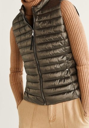 Kobieta nosząca ciemnooliwkową pikowaną bezrękawnik na beżowym prążkowanym swetrze z golfem oraz beżowe spodnie z wysokim stanem.