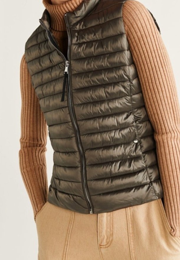 Femme portant une doudoune sans manches matelassée vert olive foncé sur un pull à col roulé côtelé couleur beige clair et un pantalon beige taille haute.