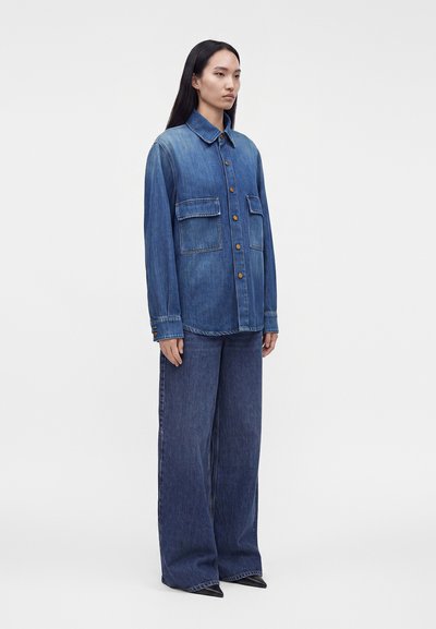 Femme portant une chemise en jean bleue à boutons avec de grandes poches et un jean bleu à jambes larges sur un fond blanc uni.