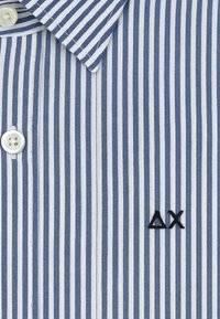 Camicia a righe blu e bianche con colletto a punta, bottoni bianchi e un logo nero ricamato sul lato sinistro. Tessuto in cotone.