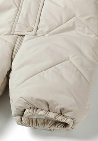 Jachetă bej, captusită, cu un model de matlasare în formă de diamant, închidere cu fermoar și manșete elastice pentru o ajustare confortabilă. Finisaj mat, moale.