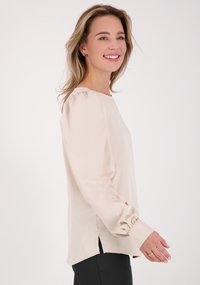 Beige blouse van zacht, glad materiaal met een ronde hals, lange poffende mouwen en zijsplitten aan de zoom voor extra bewegingsvrijheid.
