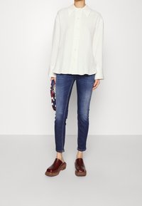 Camisa blanca de botones con un gran collar, combinada con jeans azules ajustados y zuecos marrones. La camisa tiene un corte holgado y una tela texturizada.