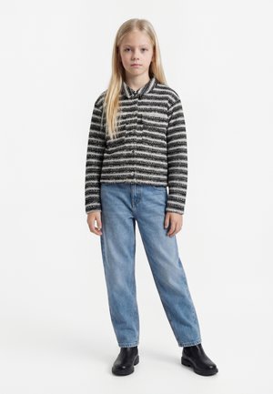 Jauna meitene ar gariem blondiem matiem melnas un baltas svītrainas jakas, brīvām zilām džinsu biksēm un melnām potīšu zābakiem, stāv baltā fonā.