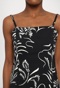 Femme portant une robe noire sans manches avec de fines bretelles et des motifs floraux abstraits blancs.