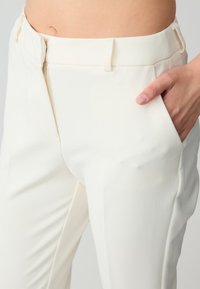 Cream-colored trousers with a tailored fit, featuring a zip closure, side pockets, and a smooth, textured fabric.