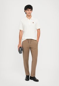 Hvid polo med et lille logo, parret med brune strækbukser. Modellen holder en grå accessoireset og har sorte sko på.