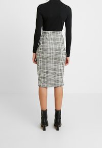 Person som bär en svart långärmad topp, hög midjerutad grå och vit blyertskjol, samt svarta ankelstövlar, står mot en vit bakgrund.