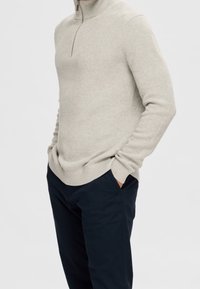 Hellgrauer Strickpullover mit halbem Reißverschluss, der einen gerippten Kragen und Bündchen aufweist, kombiniert mit marineblauen Hosen, die eine legere Passform und eine strukturierte Materialqualität betonen.