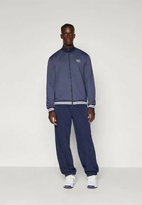 Chándal deportivo en color azul marino que consiste en una chaqueta con cremallera y pantalones a juego. La chaqueta tiene puños a rayas blancas y un logo en el pecho.