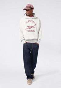 Giovane che indossa un felpa grigia "Panthers Training", jeans larghi blu scuro, scarpe ginniche beige e un cappellino rosa polveroso, in piedi con le mani in tasca.