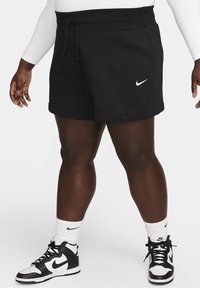 Osoba v černých Nike šortkách s bílým logem, bílých Nike ponožkách a černobílých Nike teniskách na jednobarevném bílém pozadí.