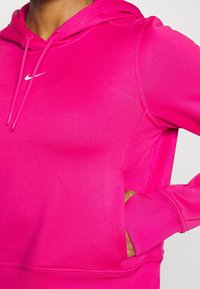 Personne portant un sweat à capuche Nike rose vif avec le logo swoosh blanc, main dans la poche avant, gros plan sur le haut du torse et le bras.