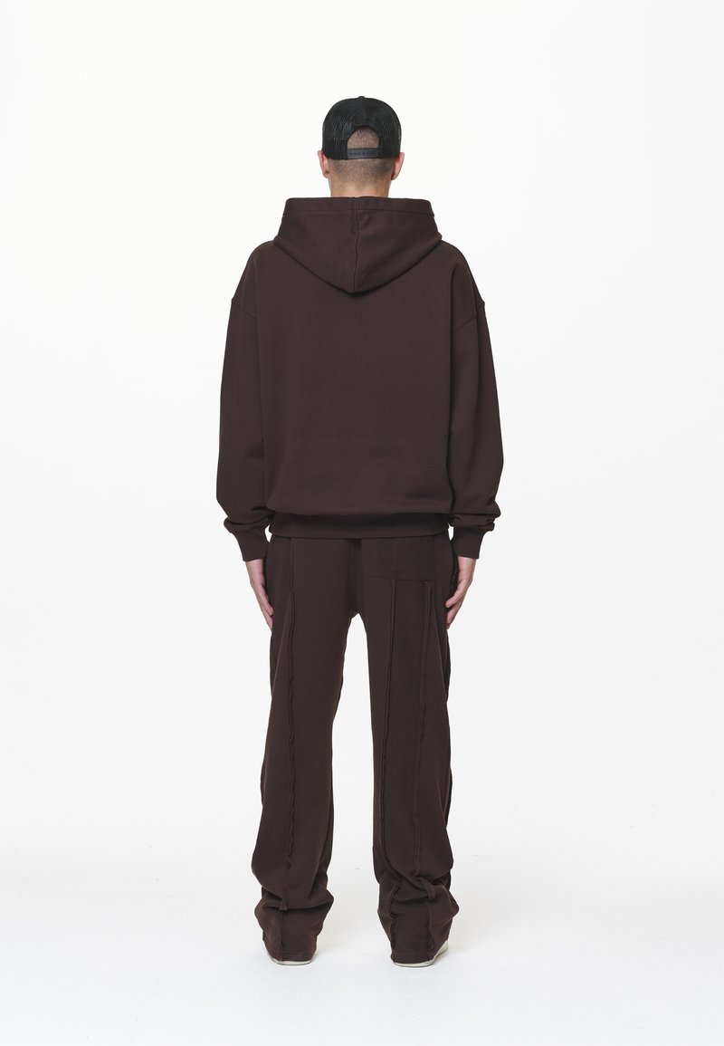 Brauner Kapuzenpullover mit großer Kapuze und großzügigem Schnitt, kombiniert mit passenden braunen Hosen, die vertikale Nähte und seitliche Taschen haben. Eine schwarze Mütze wird getragen.