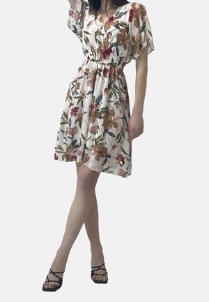 Mujer con un vestido blanco corto con estampado floral y mangas abullonadas, y sandalias negras de tiras, de pie con las piernas cruzadas.