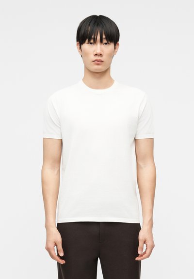Jeune homme aux cheveux noirs raides portant un t-shirt blanc uni à manches courtes et un pantalon noir, debout devant un fond gris clair uni.