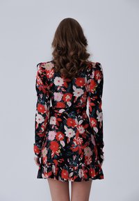 Mujer con largo cabello rizado castaño, vestida con un vestido negro de flores con flores rojas, blancas y rosas, de pie de espaldas contra un fondo liso.