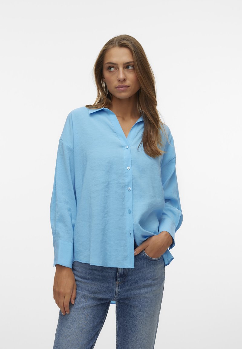 Camicia azzurro chiaro con colletto, maniche lunghe e vestibilità morbida, abbinata a jeans blu. Il tessuto sembra morbido e leggermente texturizzato.
