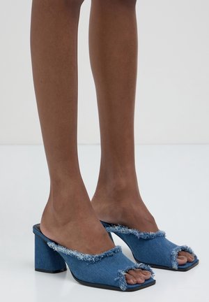 Denim-Mules mit Blockabsatz, ausgefransten Kanten und offenem Zehenbereich. Verfügt über eine abgerundete Form und ein strukturiertes Obermaterial in Blau, kontrastierender schwarzer Sohle.