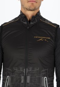 Schwarze Weste mit geometrischen grauen Mustern, Reißverschluss und dem goldenen "SWEDEMOUNT"-Logo. Mit hohem Kragen und zwei Seitentaschen.