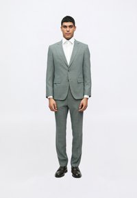 Gaišs pelēks uzvalks ar smalku tekstūru, ar iegrieztām atlokiem, divām priekšējām kabatām un vienu pogu aizdari. Kombinēts ar baltu krekla un melnām apaviem.